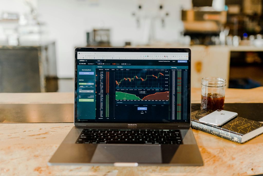 Laptop displaying stock trading dashboard, with a book and iced coffee beside it — representing an investment learning environment.